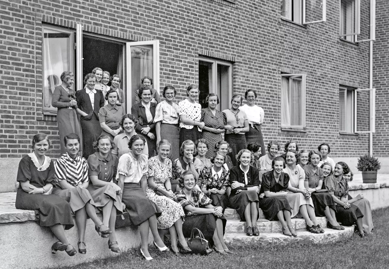 Många unga kvinnor sitter framför ett tegelhus. Foto. 